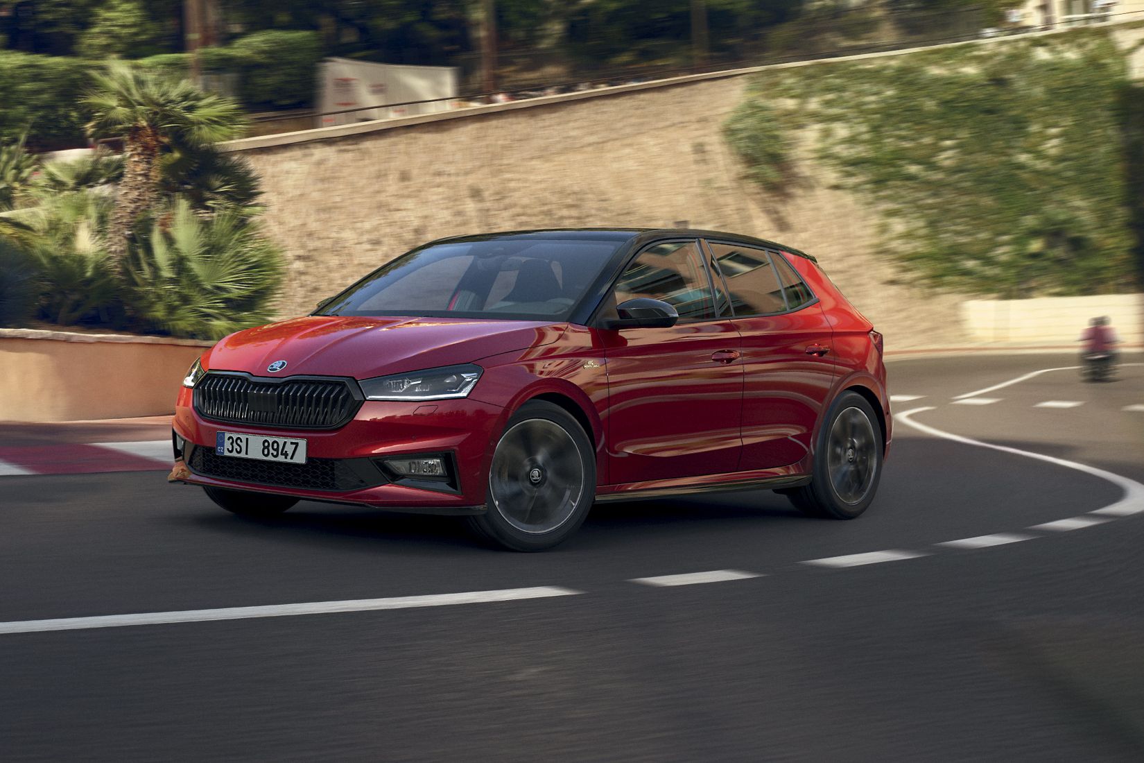 Rdeča Fabia Monte Carlo avtomobil se vozi po vijugasti cesti, obdani z zelenjem in kamnitimi zidovi. V ozadju je motociklist. Scena je postavljena v sončnem vremenu.